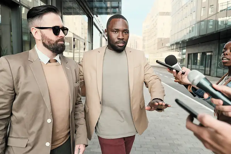 Two men in suits navigating a busy street, one gesturing as they approach a group of reporters with microphones, reflecting executive protection and security context.
